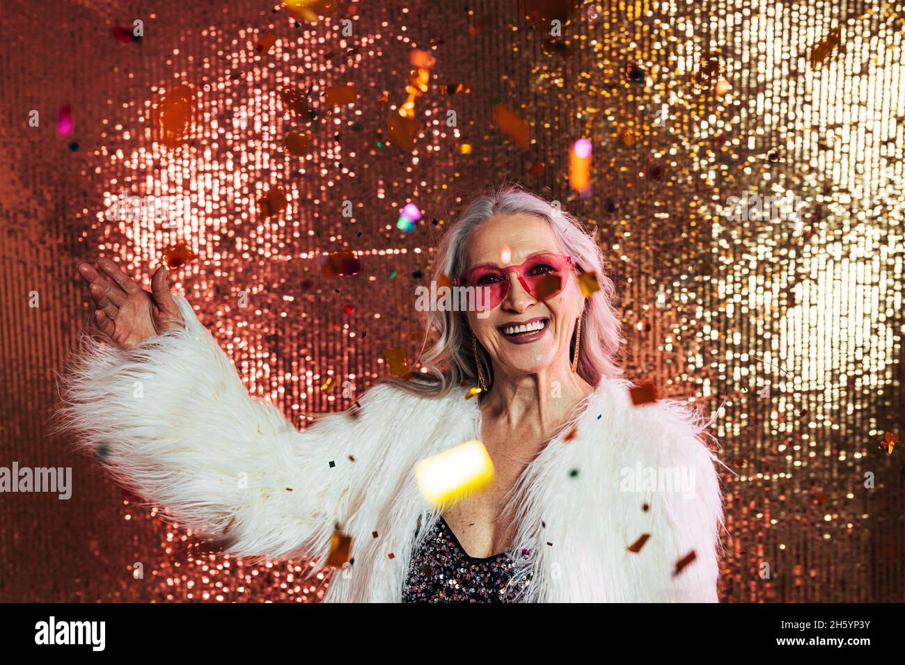 Donna anziana felice in occhiali rosa e cappotto bianco in pelliccia che si diverte sotto i confetti Foto Stock