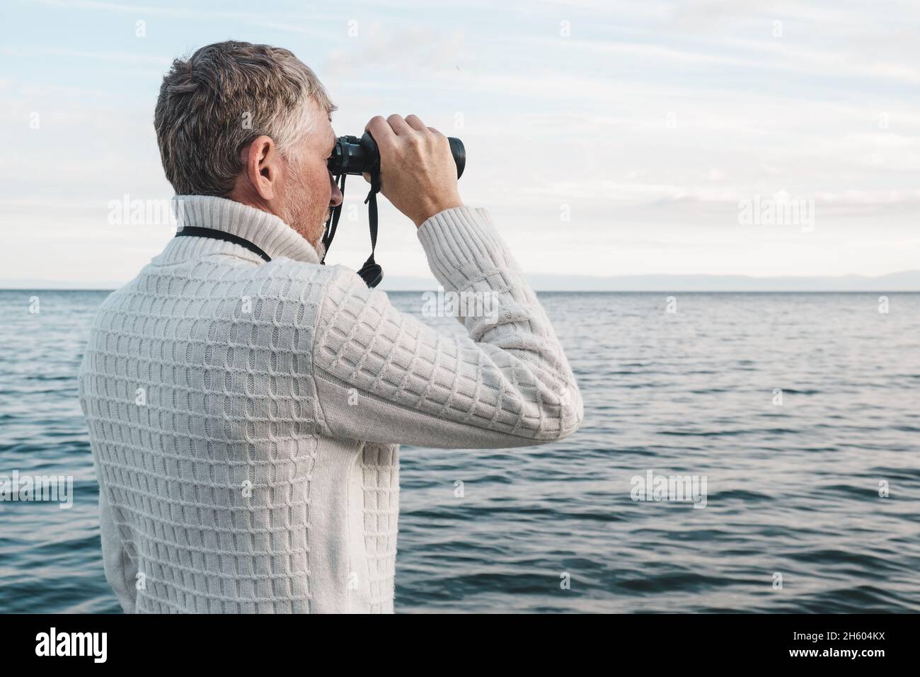 Ritratto di un bell'uomo di mezza età in un maglione bianco che guarda attraverso binocoli al mare. Spazio di copia. Foto Stock