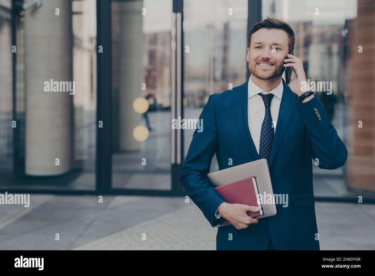 Sorridente dipendente aziendale maschile che lascia l'edificio dell'ufficio mentre parla al telefono con il collega Foto Stock