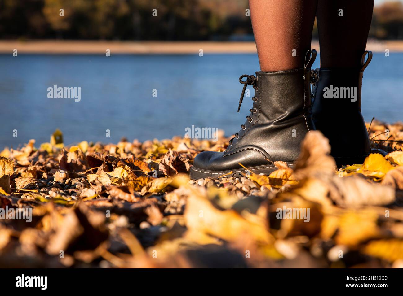 Gambe di donne in pelle nera stivali in piedi sulla spiaggia ricoperta in autunno foglie con lago sullo sfondo Foto Stock