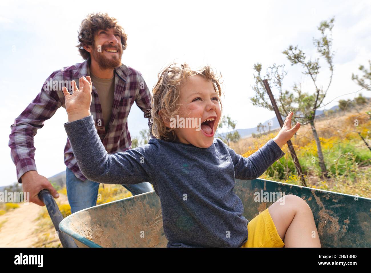 Il giovane uomo felice spinge il figlio eccitato gridando mentre si siede in carriola in fattoria Foto Stock