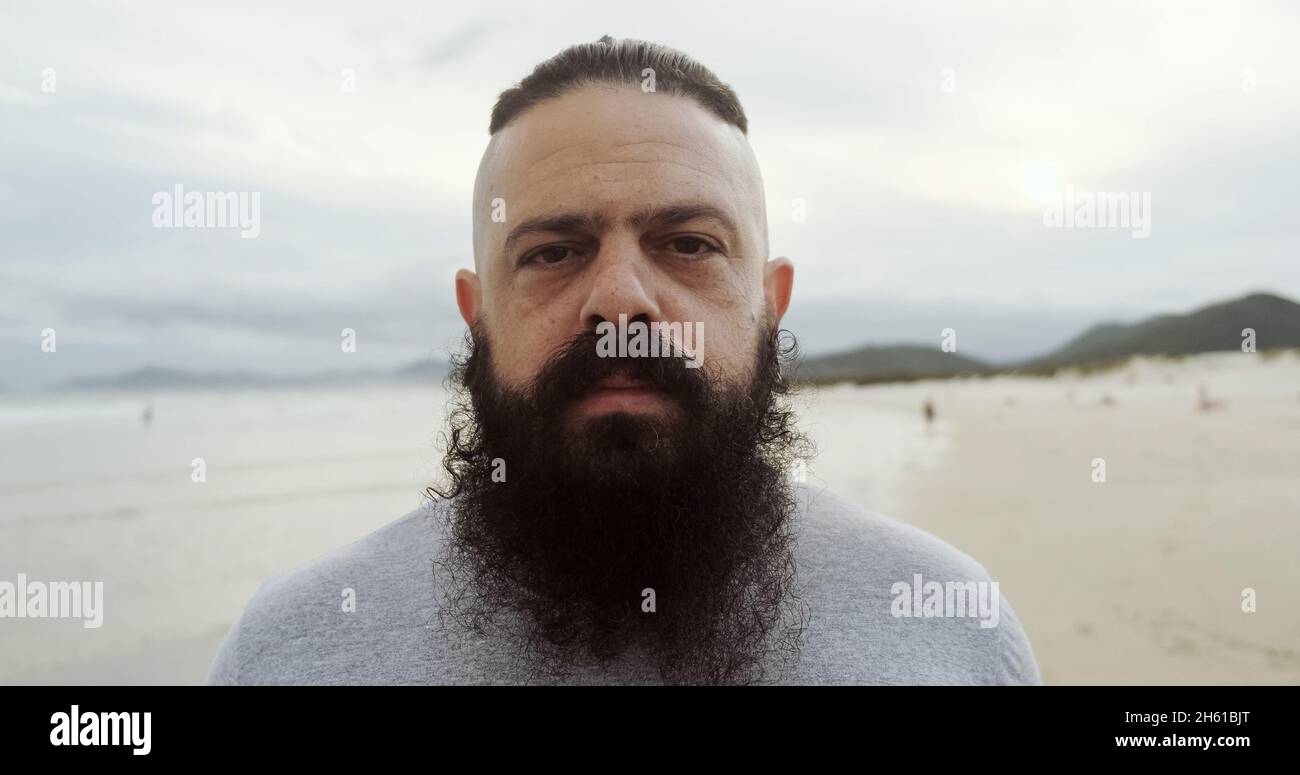 Uomo latino serio con la barba grande su una spiaggia che guarda la macchina fotografica. Foto Stock