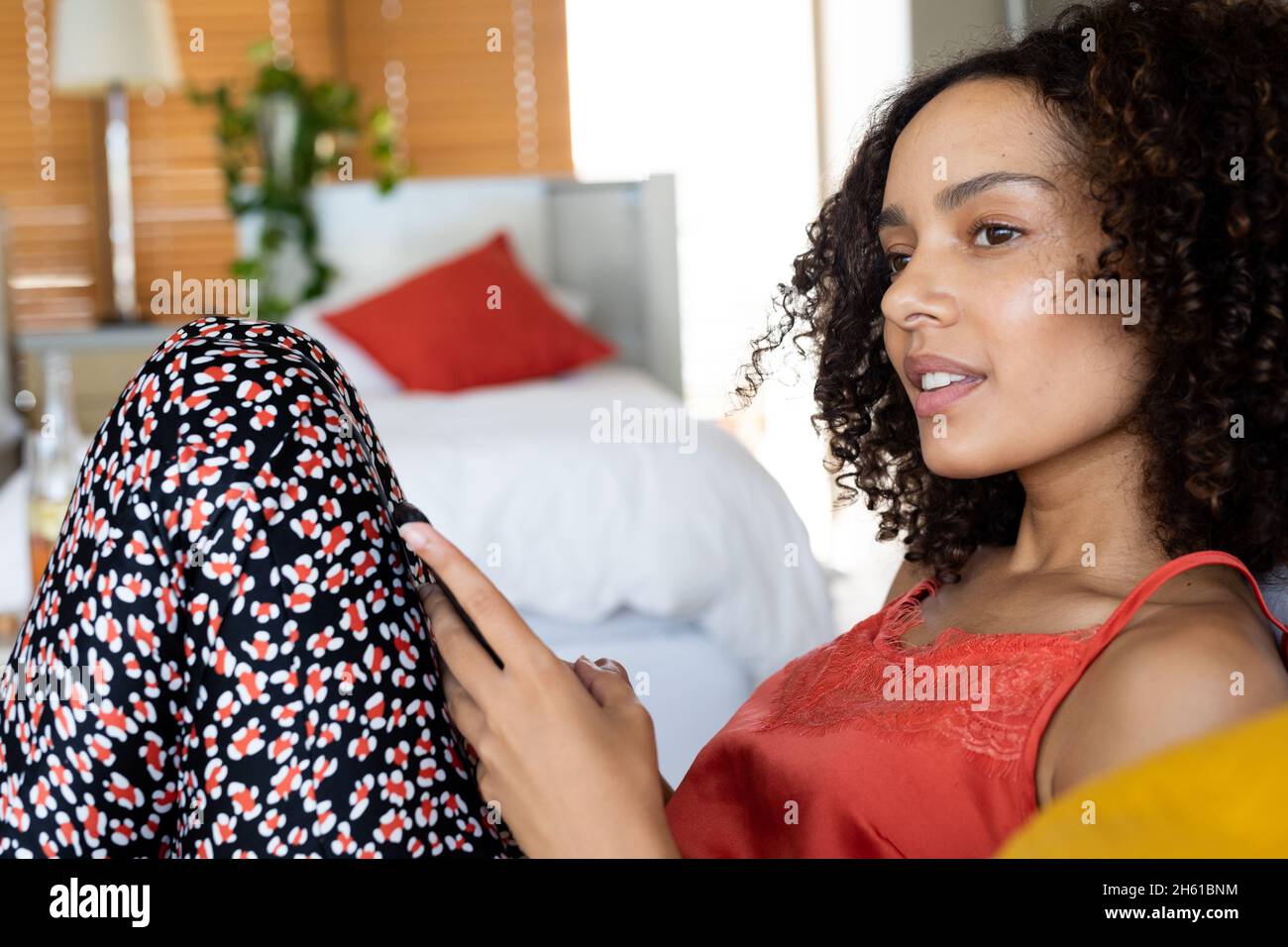 Giovane donna biraciale pensata in casuals guardando lontano mentre tiene lo smartphone a casa Foto Stock