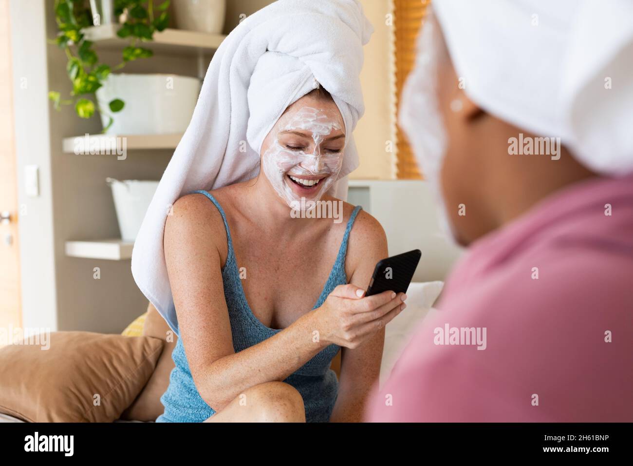 Donna felice con maschera facciale utilizzando smartphone mentre si siede con un amico a casa Foto Stock