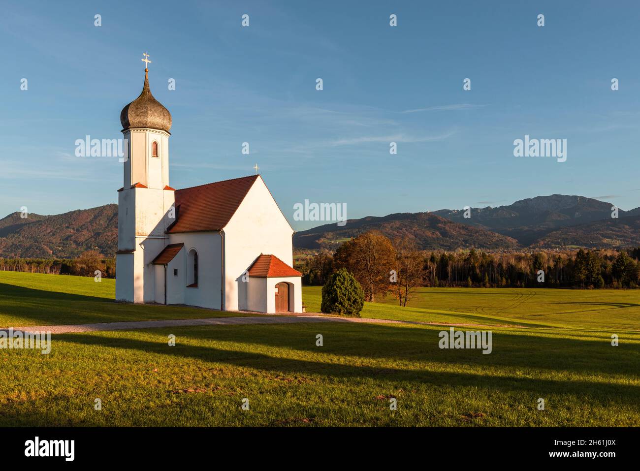 La Cappella Barocca di San Giovanni sopra la Valle di Loisach e le Prealpi autunnali bavaresi con il Monte Benediktenwand al sole serale, in Germania Foto Stock