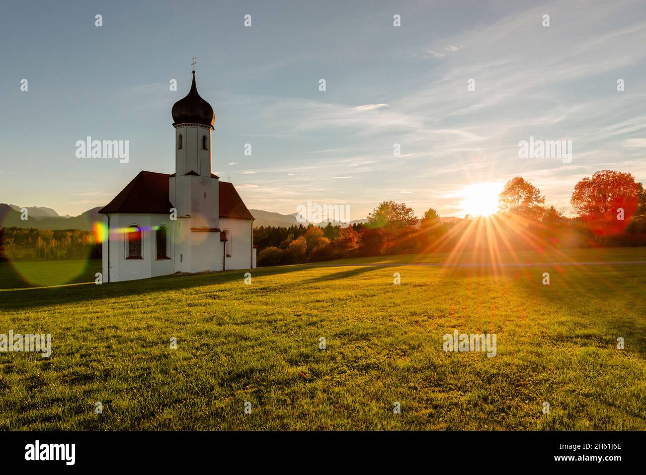 La cappella barocca di San Giovanni sopra la valle di Loisach e le Alpi bavaresi autunnali con Zugspitze in controluce del sole serale, Germania Foto Stock