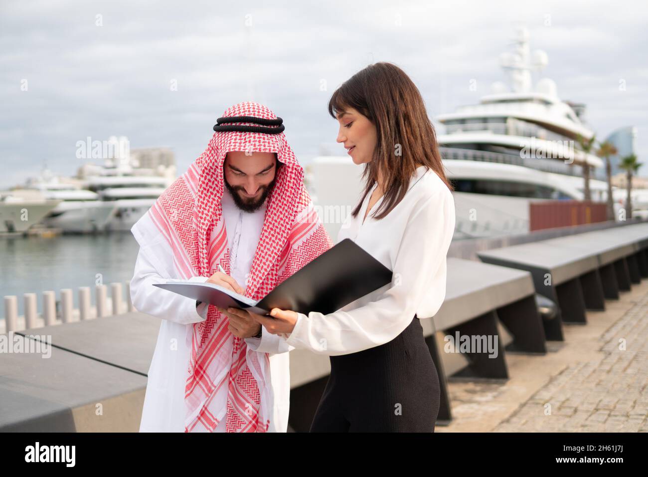 Sorridente uomo d'affari arabo che detiene contratto di canto penna mentre si trova in piedi con la giovane donna d'affari europea nel porto, uomo arabo che mette la firma sui documenti durante l'incontro all'aperto con il partner d'affari Foto Stock