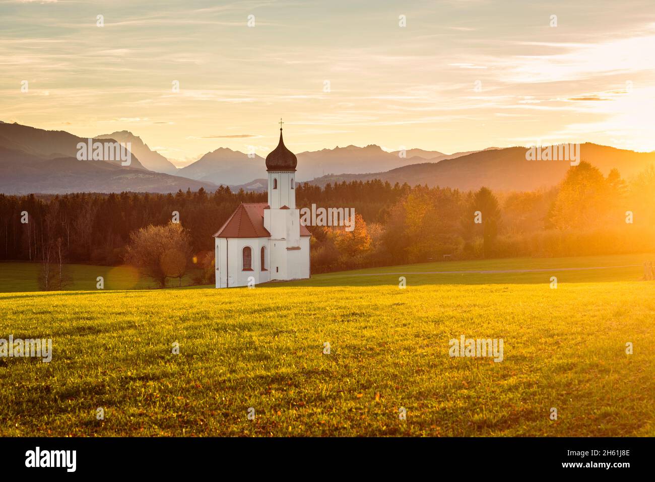 La cappella barocca di San Giovanni sopra la valle di Loisach e le Alpi bavaresi autunnali con Zugspitze in controluce del sole serale, Germania Foto Stock