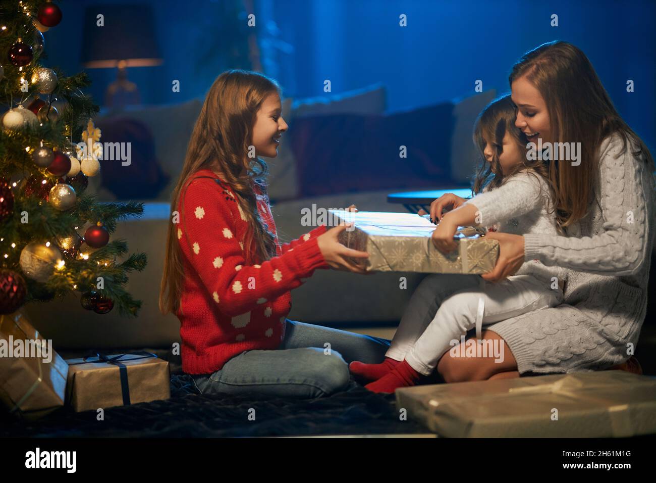 Bella donna caucasica e due bambine che aprono regali di natale sotto il bellissimo albero di natale a casa. Famiglia felice godersi le vacanze invernali. Concetto di celebrazione e tradizione. Foto Stock