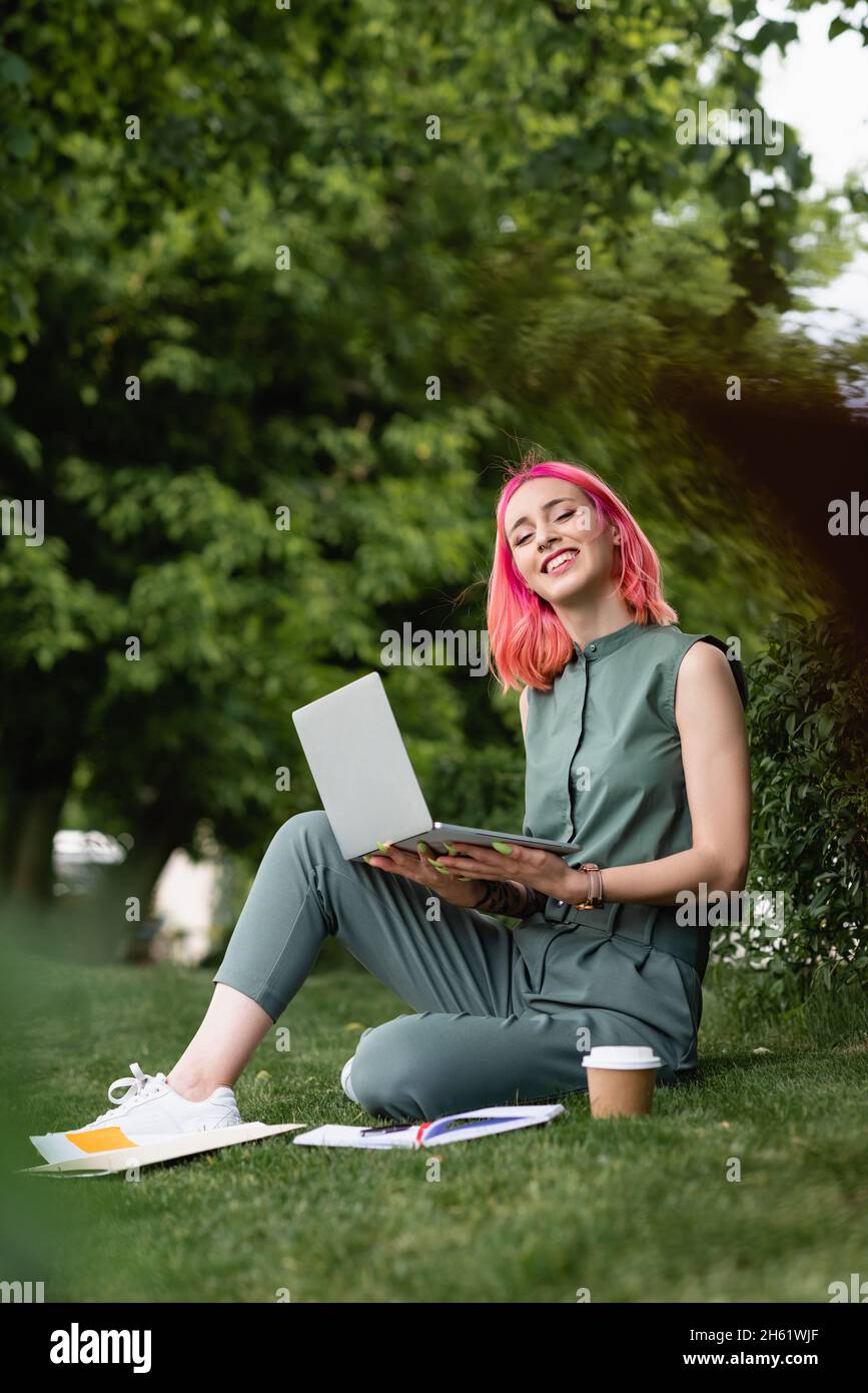 donna gioiosa con i capelli rosa che tengono il computer portatile e seduto sul prato Foto Stock
