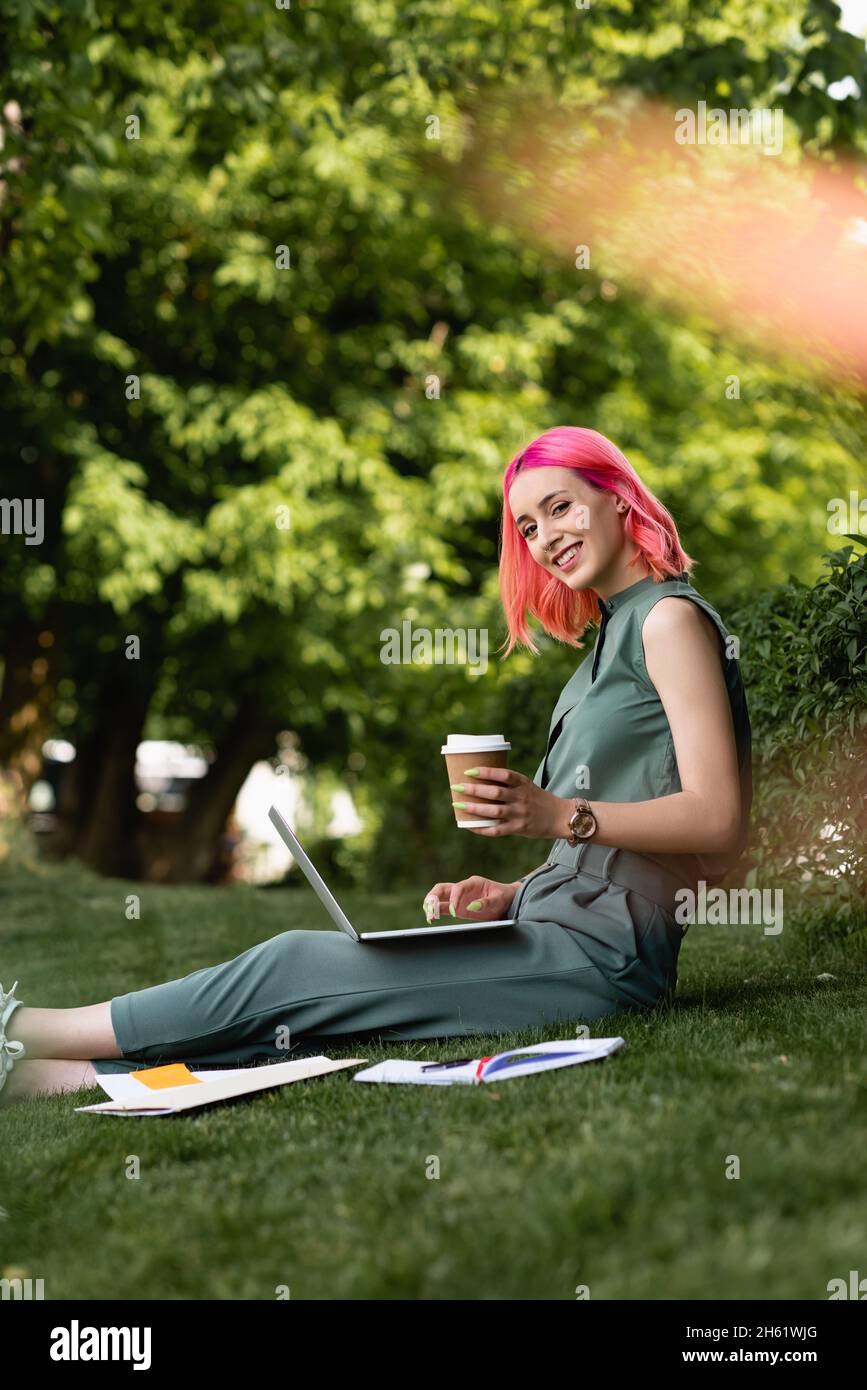 donna felice con i capelli rosa che tiene la tazza di carta e usando il laptop sull'erba Foto Stock