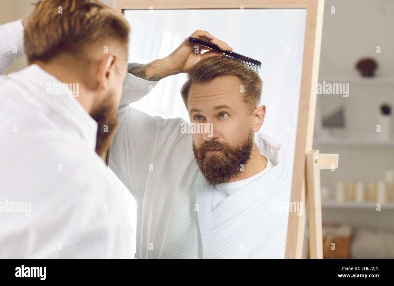Sicuro bel bearded giovane uomo guardante nello specchio del bagno e spazzolando i suoi capelli Foto Stock