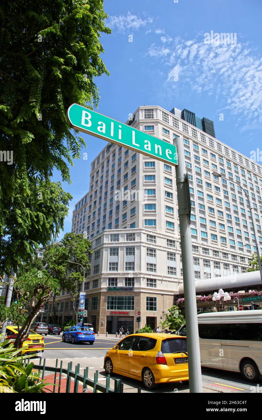 Un cartello con il nome della strada per Bali Lane con il Raffles Hospital sullo sfondo nel quartiere Bugis di Singapore. Foto Stock