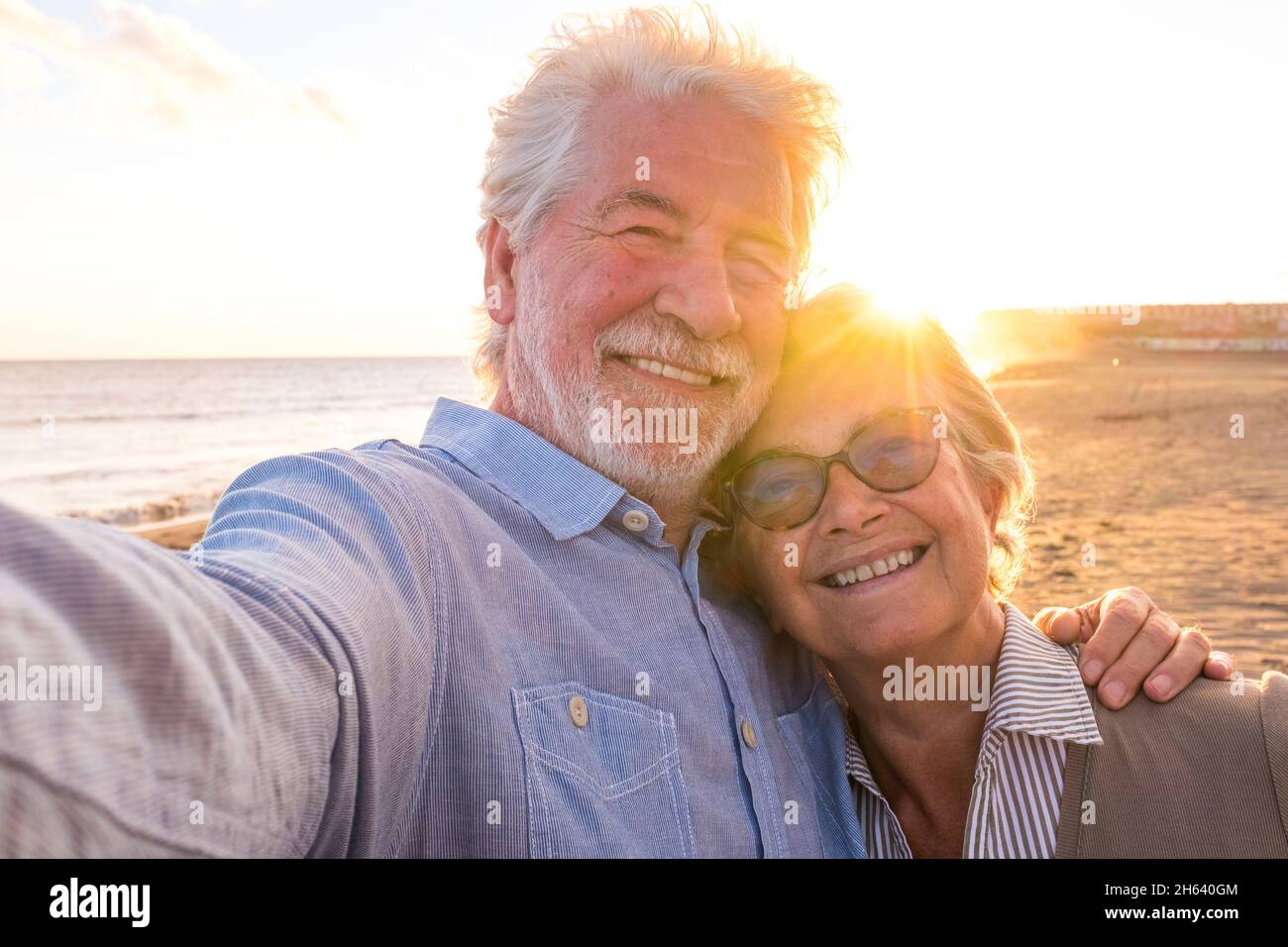 ritratto di coppia di persone anziane e mature che godono l'estate in spiaggia guardando la macchina fotografica prendendo un selfie insieme al tramonto sullo sfondo. due anziani attivi che viaggiano all'aperto. Foto Stock