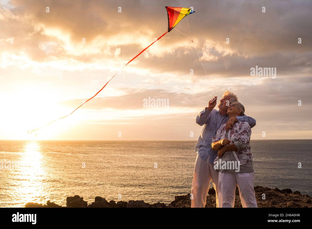 primo piano e ritratto di due persone anziane e mature giocare e divertirsi con un aquilone in spiaggia con il mare sullo sfondo con il tramonto - attivo gli anziani si divertono Foto Stock
