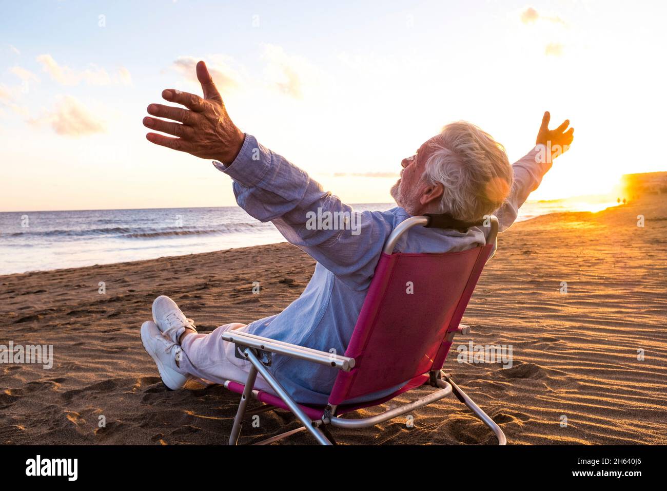 un vecchio e maturo che si gode le vacanze estive da solo sulla spiaggia seduta in una piccola sedia che guarda al mare. uomo che si sente libero con le braccia aperte. concetto di libertà. Foto Stock