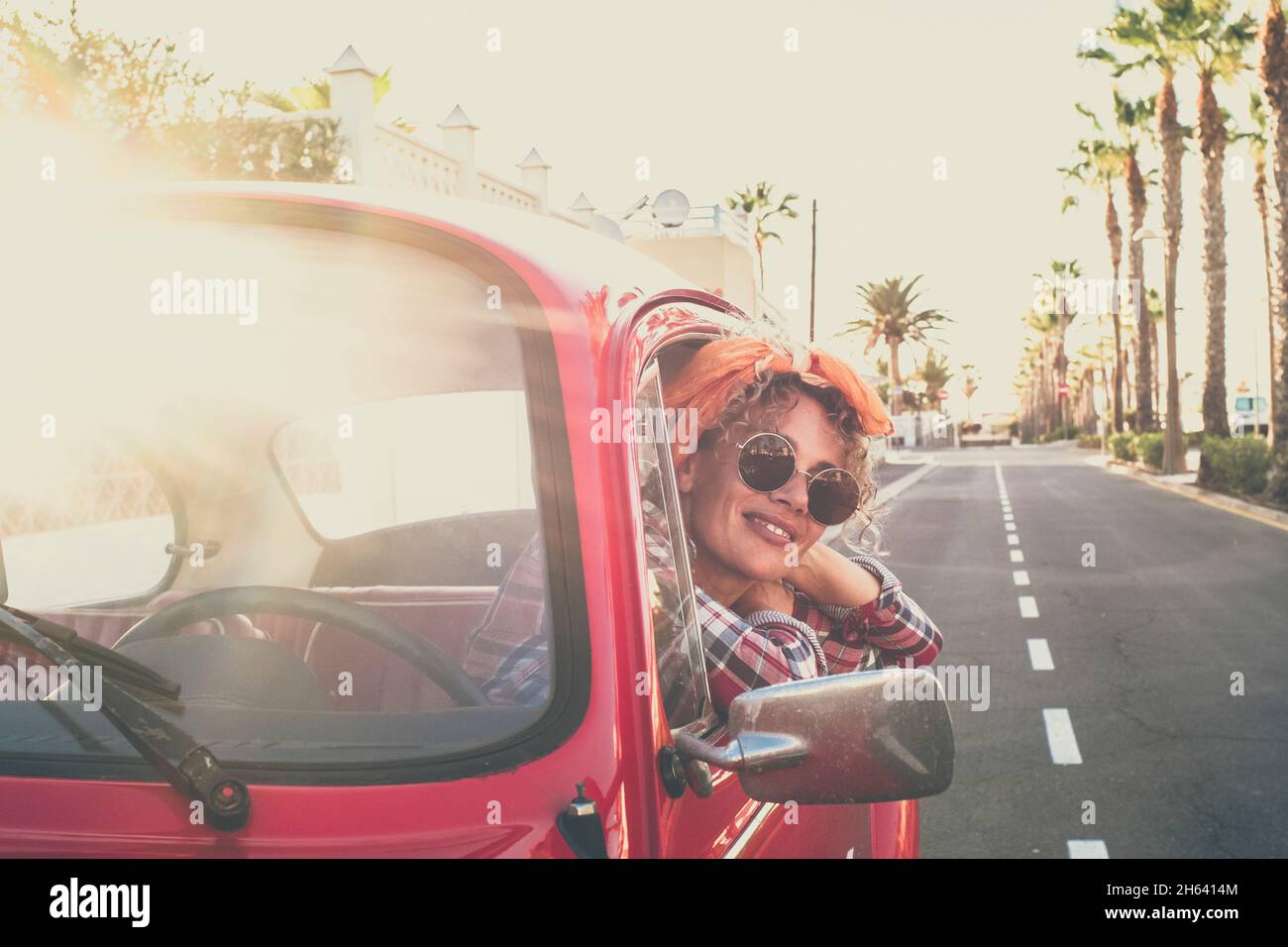 allegro– bella adulto giovane donna caucasica sorridere e godere di viaggio in piedi fuori dalla macchina rossa vecchio veicolo cercando - alla moda femminile con occhiali da sole e vestiti colorati - strada in background Foto Stock