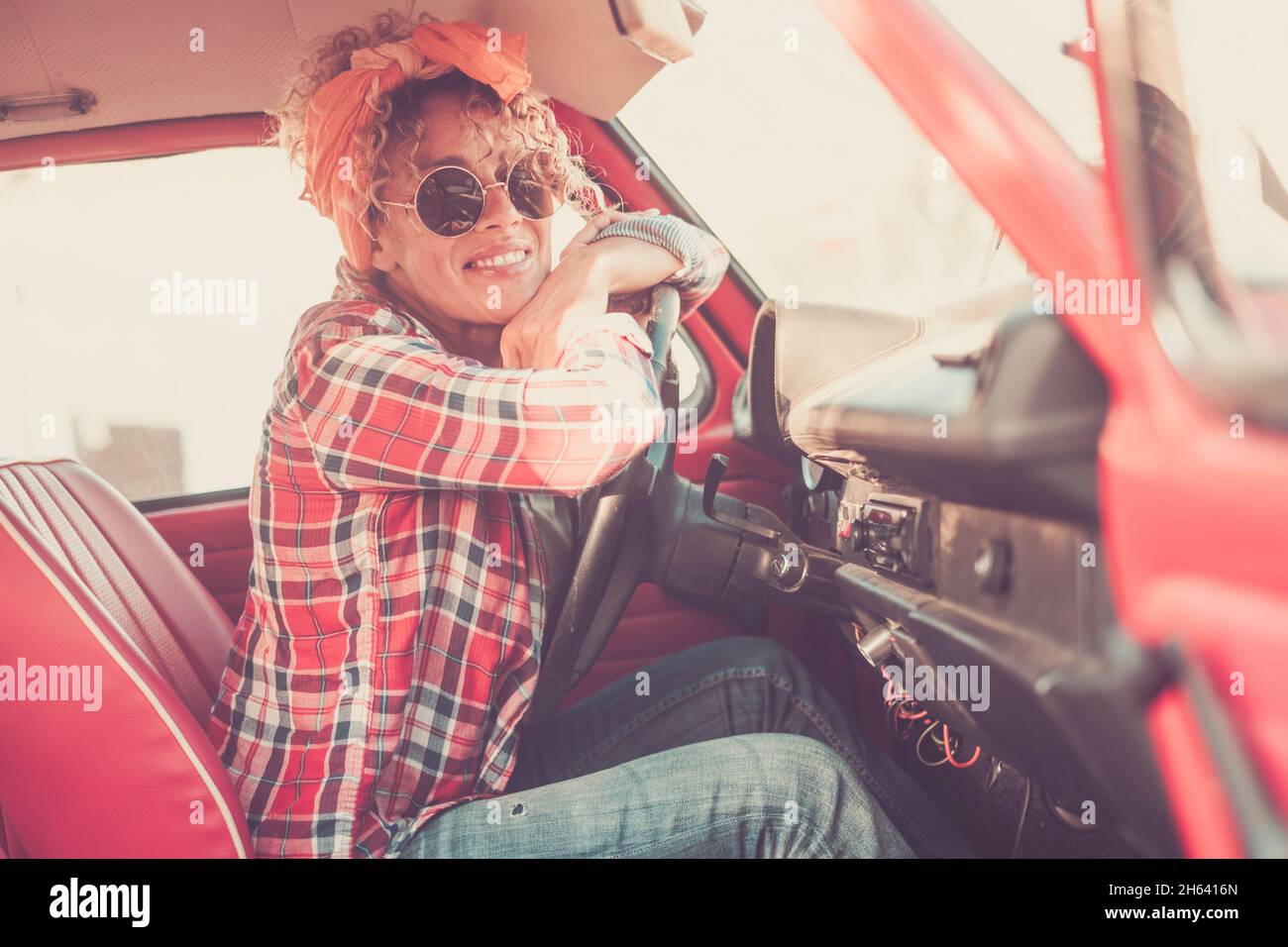 bel ritratto di giovane adulto caucasico donna di mezza età seduto all'interno di auto rosso vecchio veicolo e sorridente alla macchina fotografica indossando occhiali da sole - gente alla moda e concetto di trasporto Foto Stock
