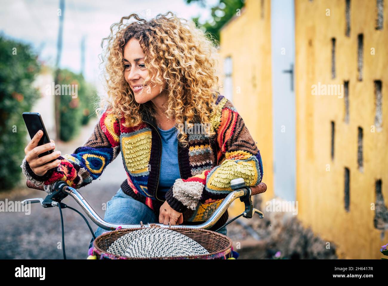felice bella adulta giovane donna godere di attività di svago all'aperto su una bicicletta e leggere il telefono cellulare sorridente - la gente di donna usa cellulare moderno per inviare il messaggio - bionda capelli ricci lungo allegro ragazza di mezza età allegra Foto Stock