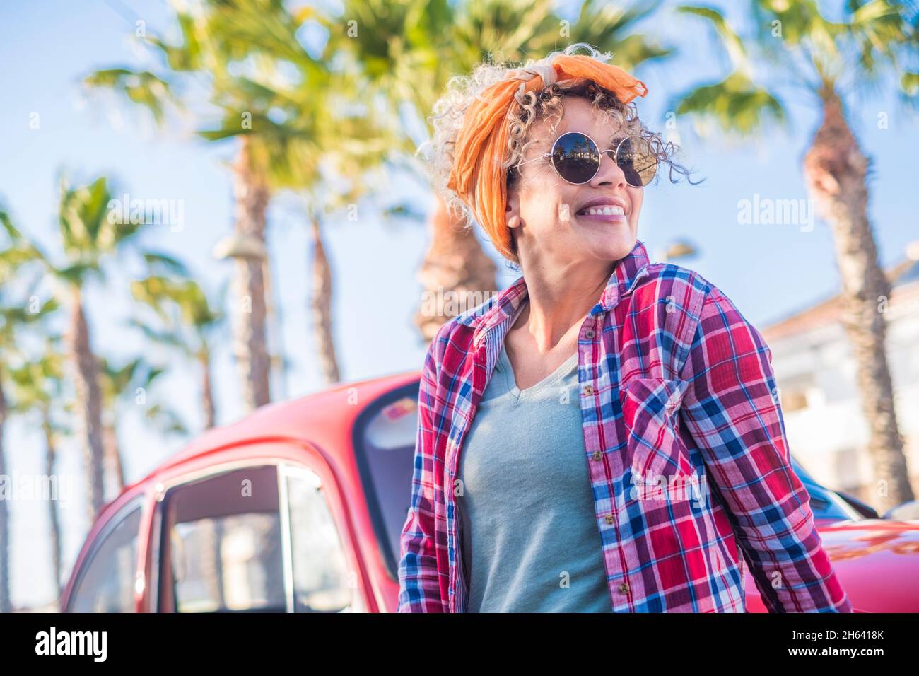 ritratto di allegro grazioso adulto giovane donna caucasica sorriso e godere il tempo di estate in piedi al di fuori della macchina rossa vecchio veicolo guardando - alla moda femminile persone con occhiali da sole e vestiti colorati Foto Stock