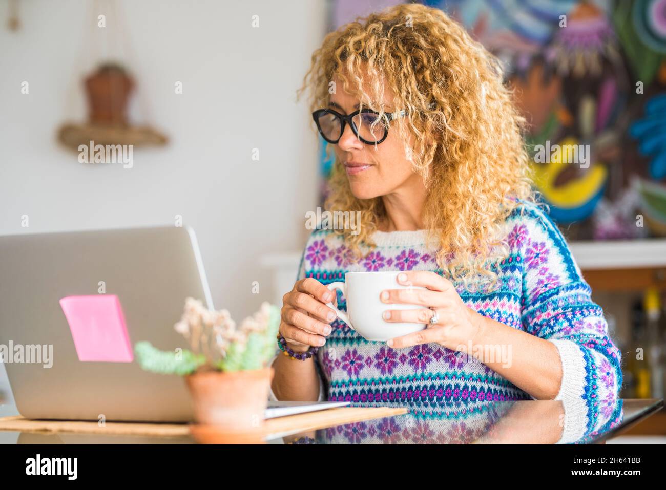 donna caucasica adulta guardare computer portatile a casa bere con una tazza - piuttosto bionda donne persone utilizzare la tecnologia per il lavoro o il tempo libero - collega giovane donna con occhiali neri Foto Stock