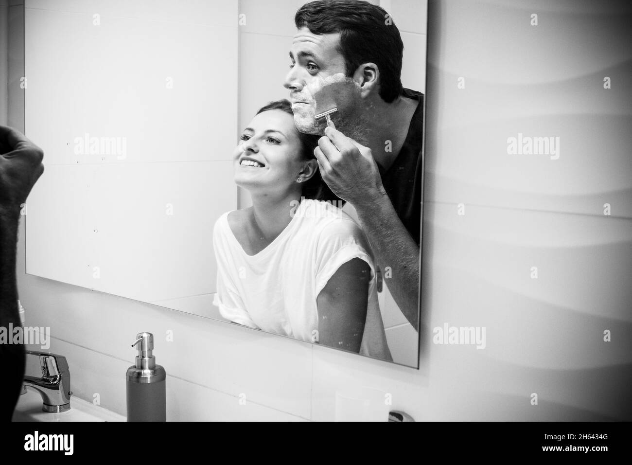 coppia gioiosa che si diverte in bagno durante la mattina mentre l'uomo sta rasando la barba usando il rasoio e la schiuma. coppia felice che si prepara insieme nel bagno domestico Foto Stock