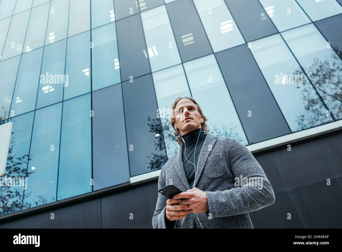 Uomo d'affari moderno con smartphone nelle sue mani su una strada della città. Bell'uomo che indossa le cuffie e parla sul telefono cellulare, ascoltando musica o podc Foto Stock