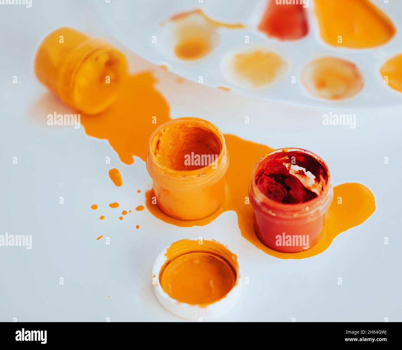 Su una tavola bianca c'è una tavolozza bianca colorata con vernice, e accanto ad essa ci sono vasi di gouache in rosso e giallo, uno dei quali è versato. Cre Foto Stock