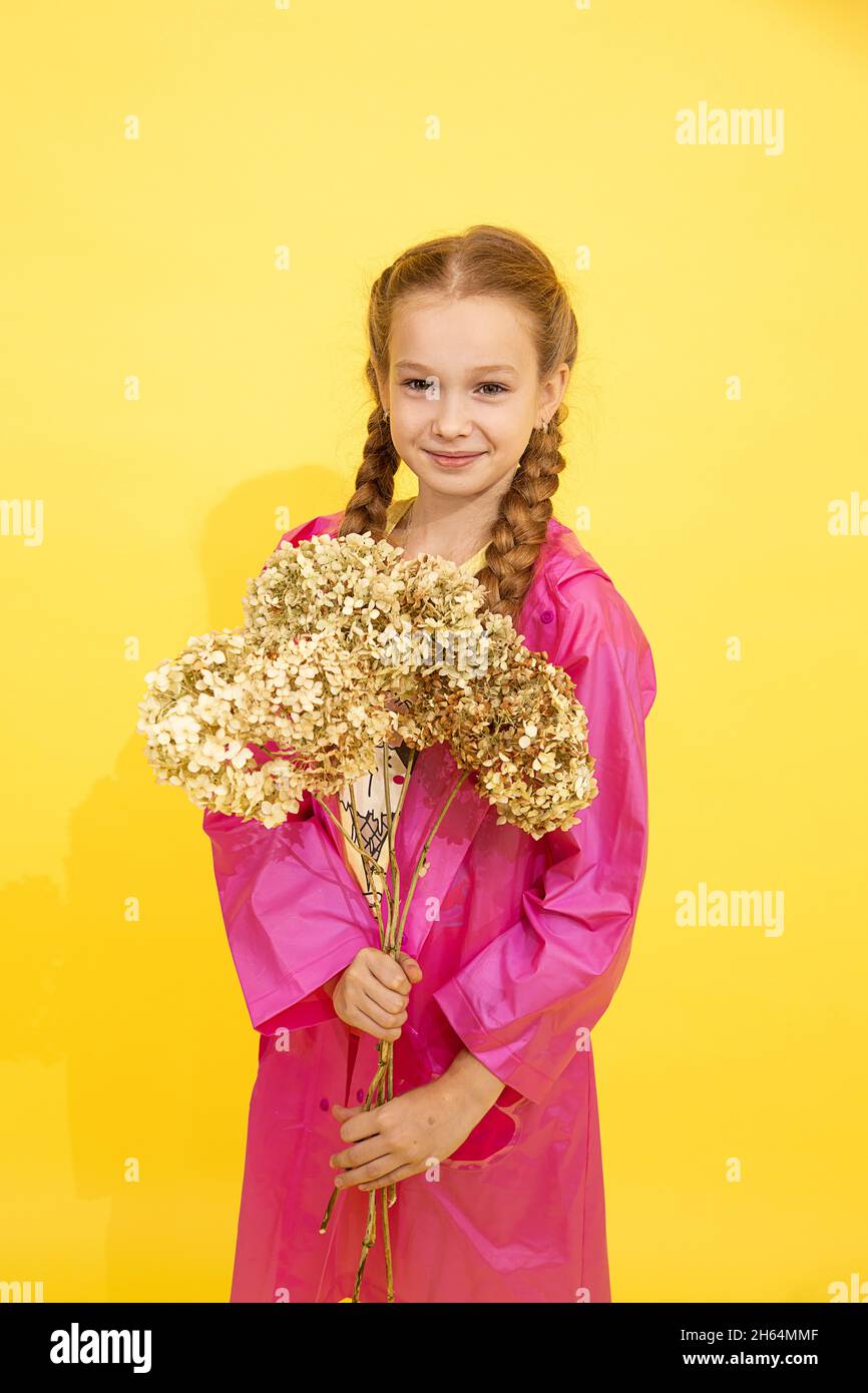 bella ragazza di 7-8 anni con due pigtail in impermeabile rosa con fiore su sfondo giallo in studio guarda la macchina fotografica e sorrisi. Festa dell'insegnante, di Madre Foto Stock