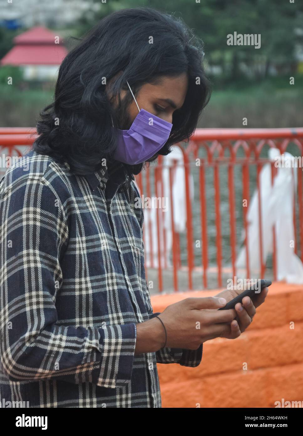 Vista laterale di un uomo giovane indiano a capelli lunghi che indossa maschera facciale con l'uso del telefono mentre si trova all'esterno Foto Stock