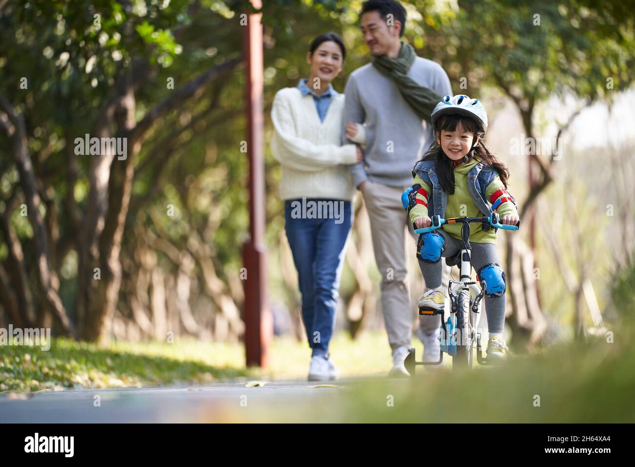 bambina asiatica con casco e completo di protezione in bicicletta nel parco della città con i genitori che guardano da dietro Foto Stock