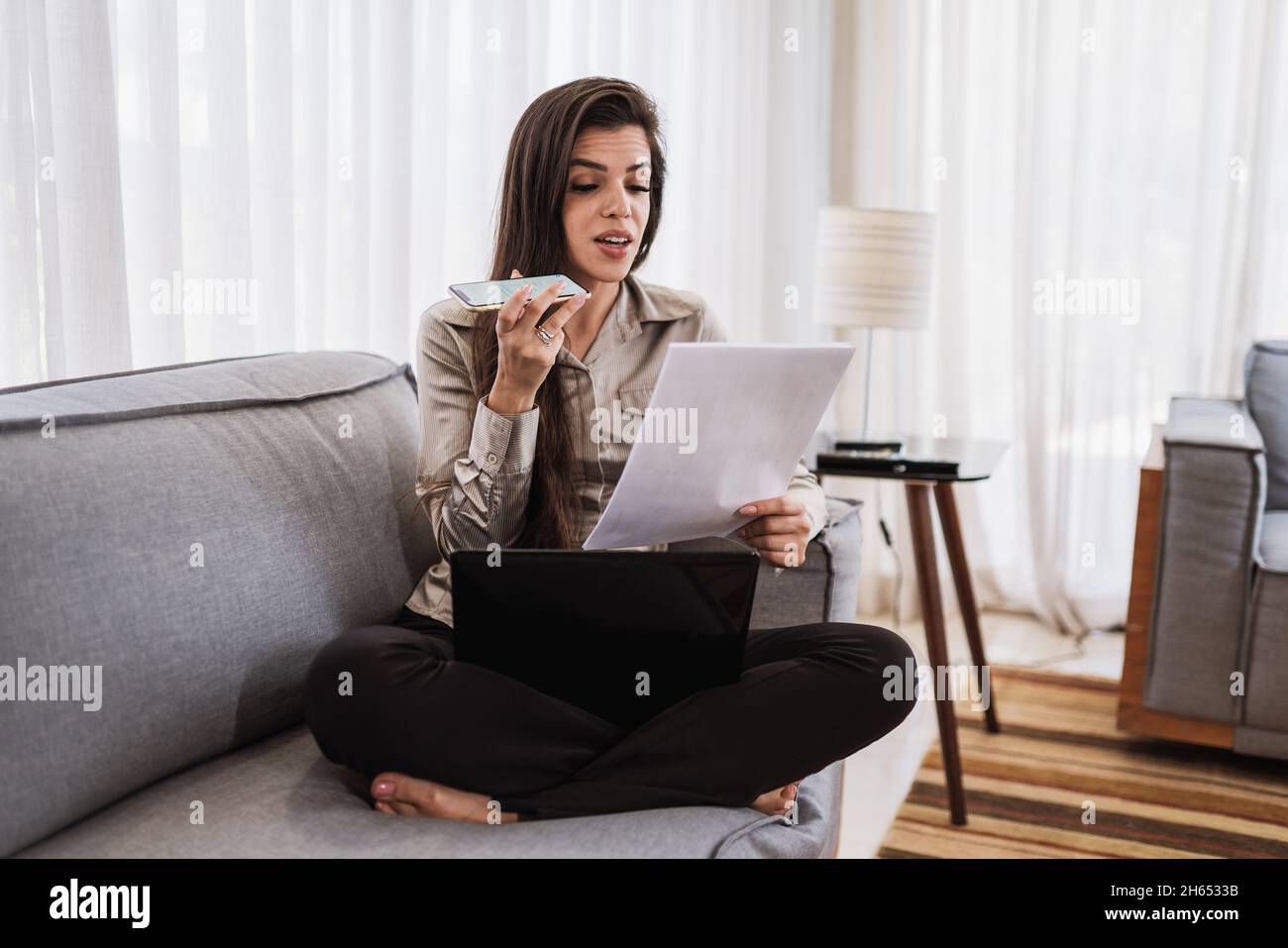 Giovane bella donna brasiliana che invia messaggi audio su smartphone e lavora con il computer portatile mentre si siede sul divano, lavorando da casa concetto. Foto Stock