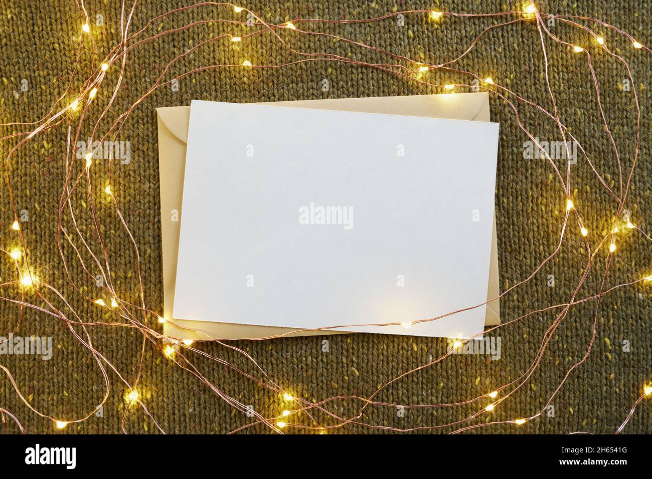 Biglietto d'auguri vuoto, busta artigianale e luci di Natale incandescente su sfondo verde lavorato a maglia Foto Stock