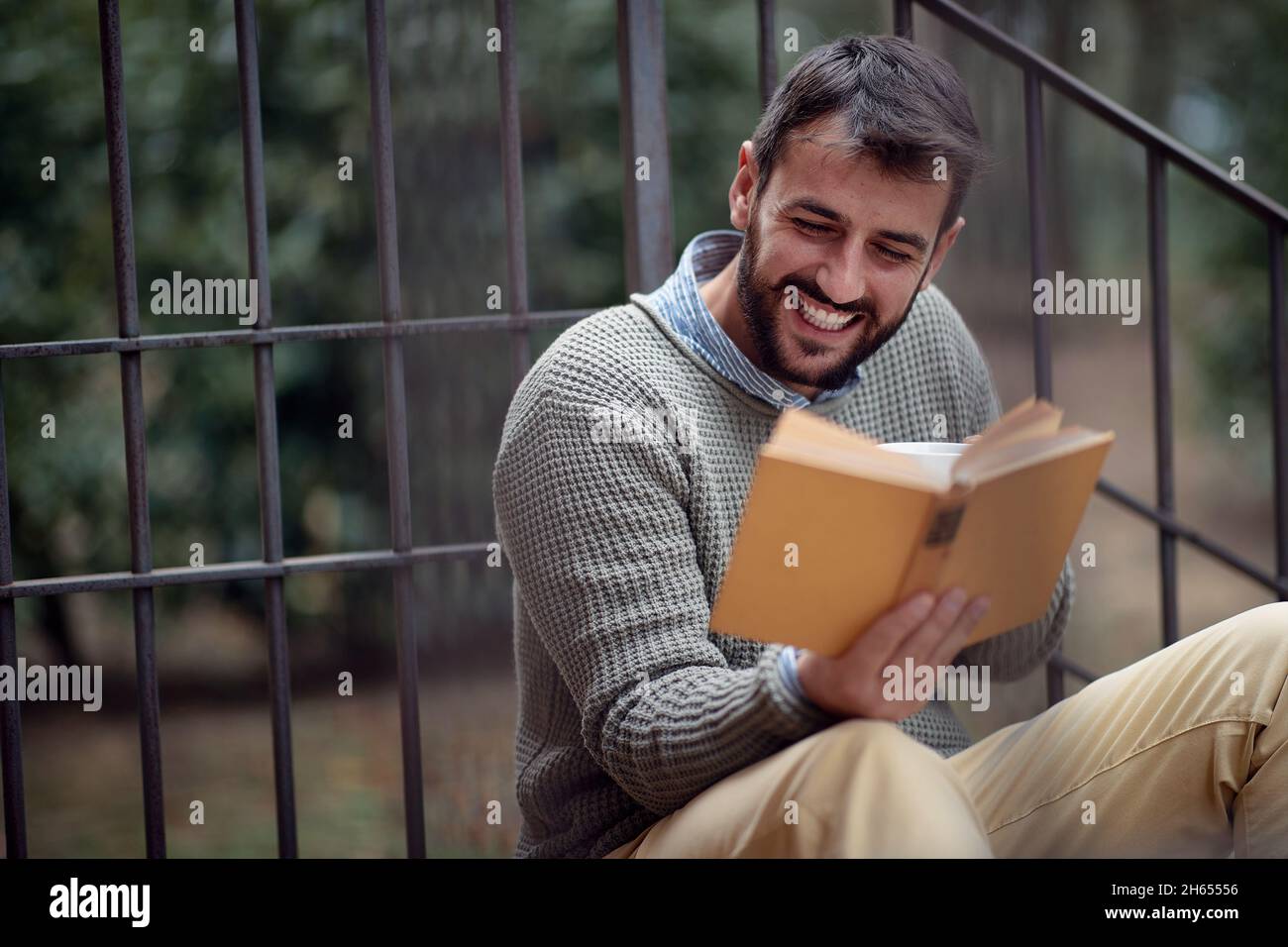 Un giovane uomo bello è felice mentre si siede sulle scale di fronte alla casa in un'atmosfera rilassata e leggendo un libro. Città, fuori, riposo Foto Stock