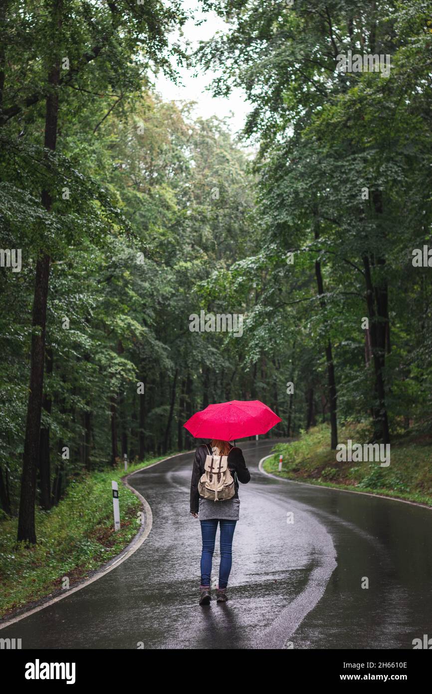 Donna viaggiatore con ombrello rosso a piedi su strada vuota nella foresta in giorno di pioggia. Backpacker viaggia da solo in cattive condizioni meteorologiche. Foto Stock
