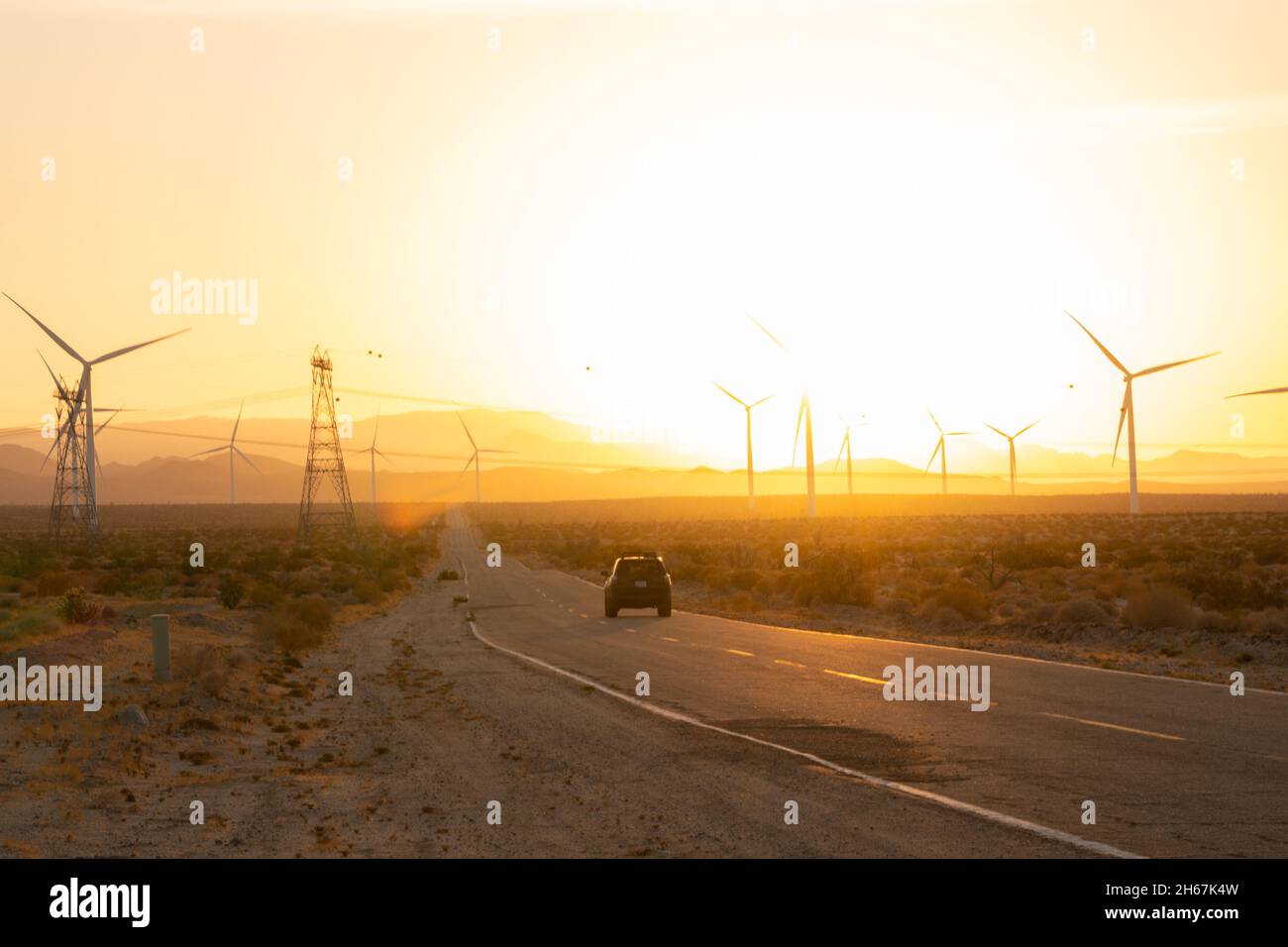 Auto che guida al tramonto con mulini a vento e piloni elettrici Foto Stock