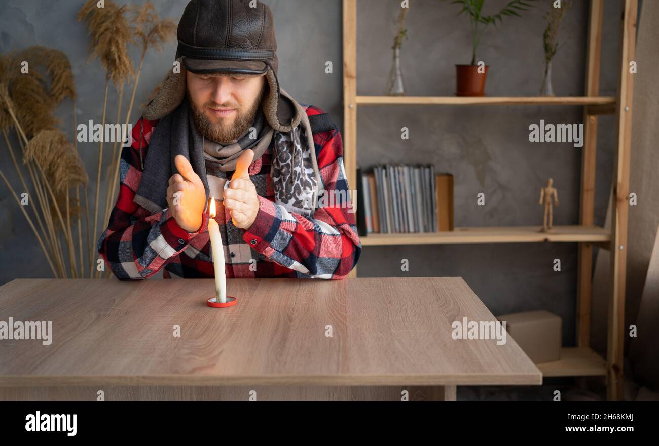 Un uomo caucasico vestito con un caldo cappello invernale e una sciarpa siede a casa a un tavolo e scalda le mani da una candela che brucia. Foto Stock