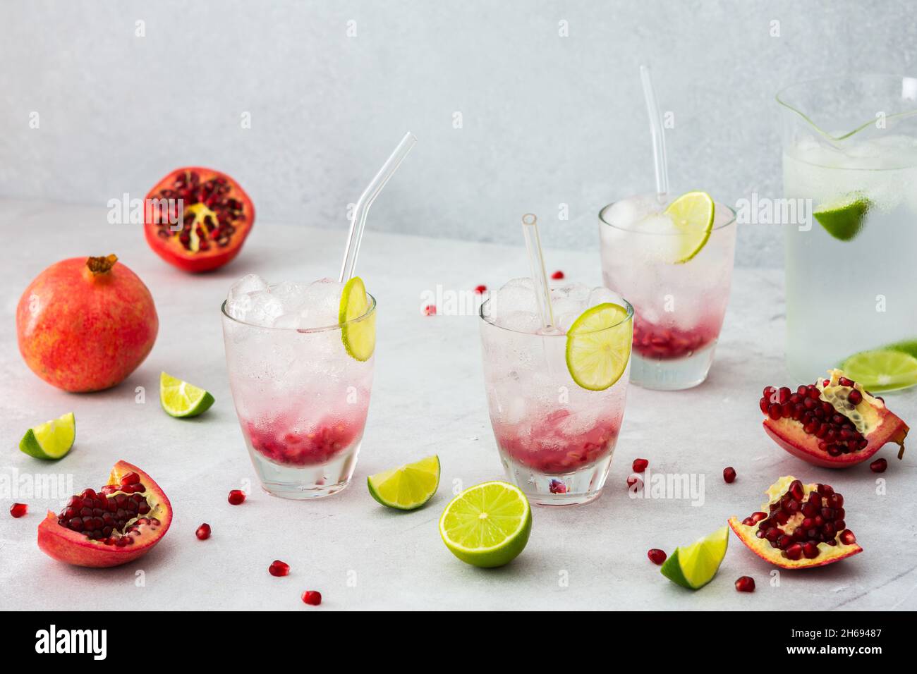 Rinfrescante bevanda di melograno in bicchieri con semi di melograno e ghiaccio, cannucce di calce e vetro, brocca di acqua fredda con agrumi, melograni tagliati Foto Stock