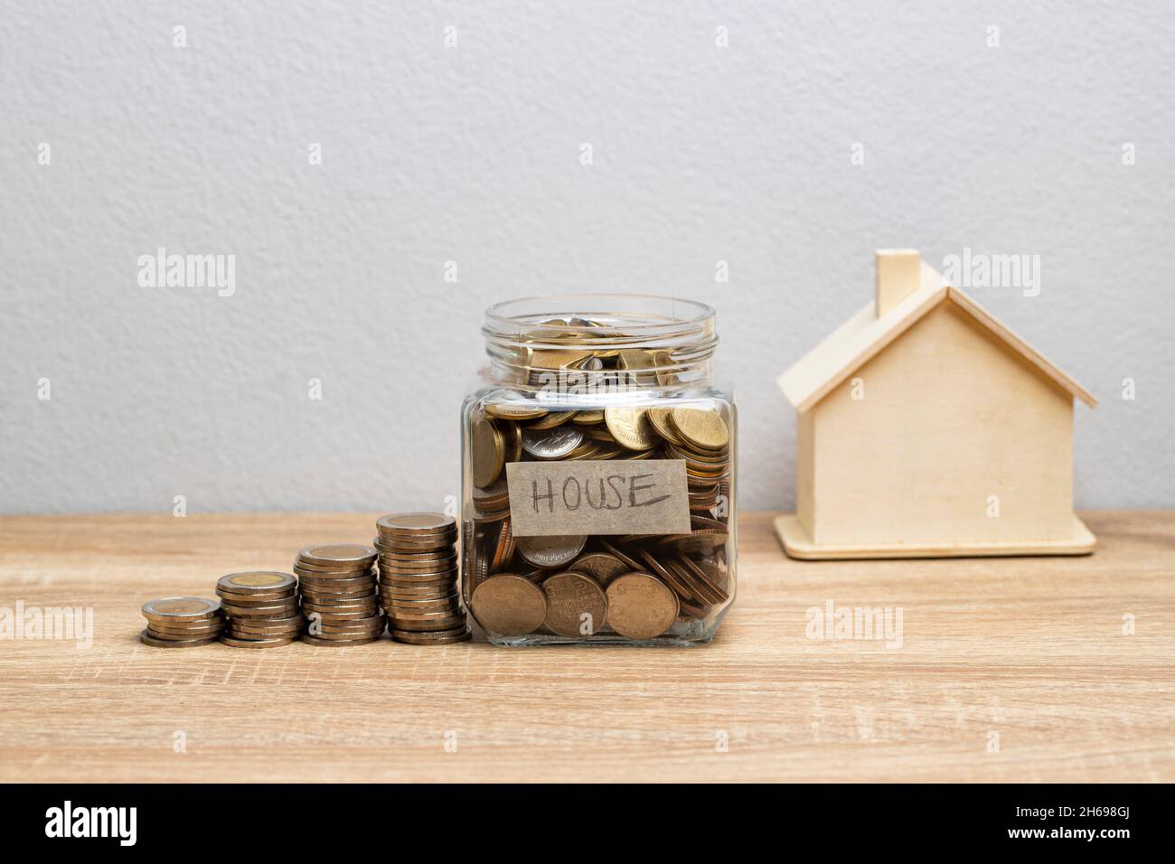 Moneta in un vaso di vetro con pila di soldi e casa di legno bianco sul tavolo marrone con spazio copia per concetto finanziario Foto Stock