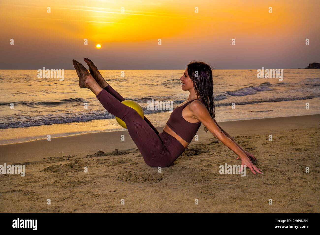 Bella donna che fa pilates esercizio con palla sulla spiaggia al tramonto. Donna seduta sulla sabbia facendo pilates esercizio tenendo palla morbida tra sollevato Foto Stock