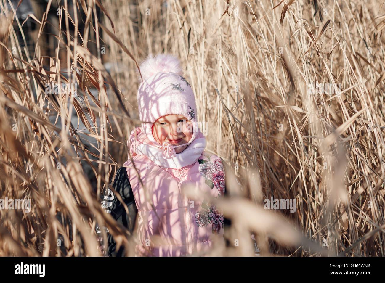 Il bambino è in piedi in erba asciutta. La bambina in abiti rosa inverno gode di sole ed esplora il thicket di canne nella giornata di sole nella natura Foto Stock