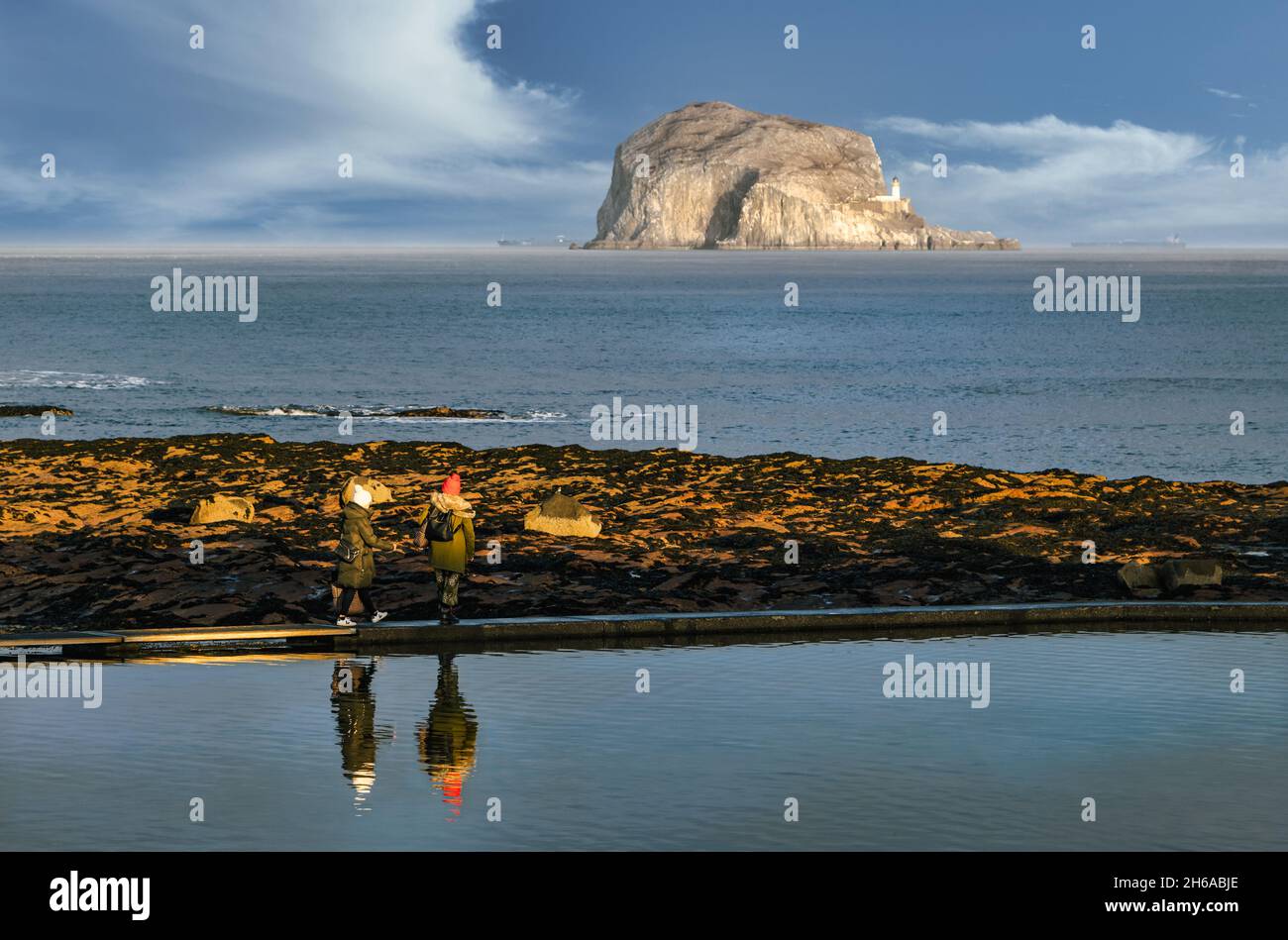 Due persone che camminano su una piscina di mare murata riflessa nell'acqua ferma con l'isola di Bass Rock in lontananza, North Berwick, Scozia, Regno Unito Foto Stock