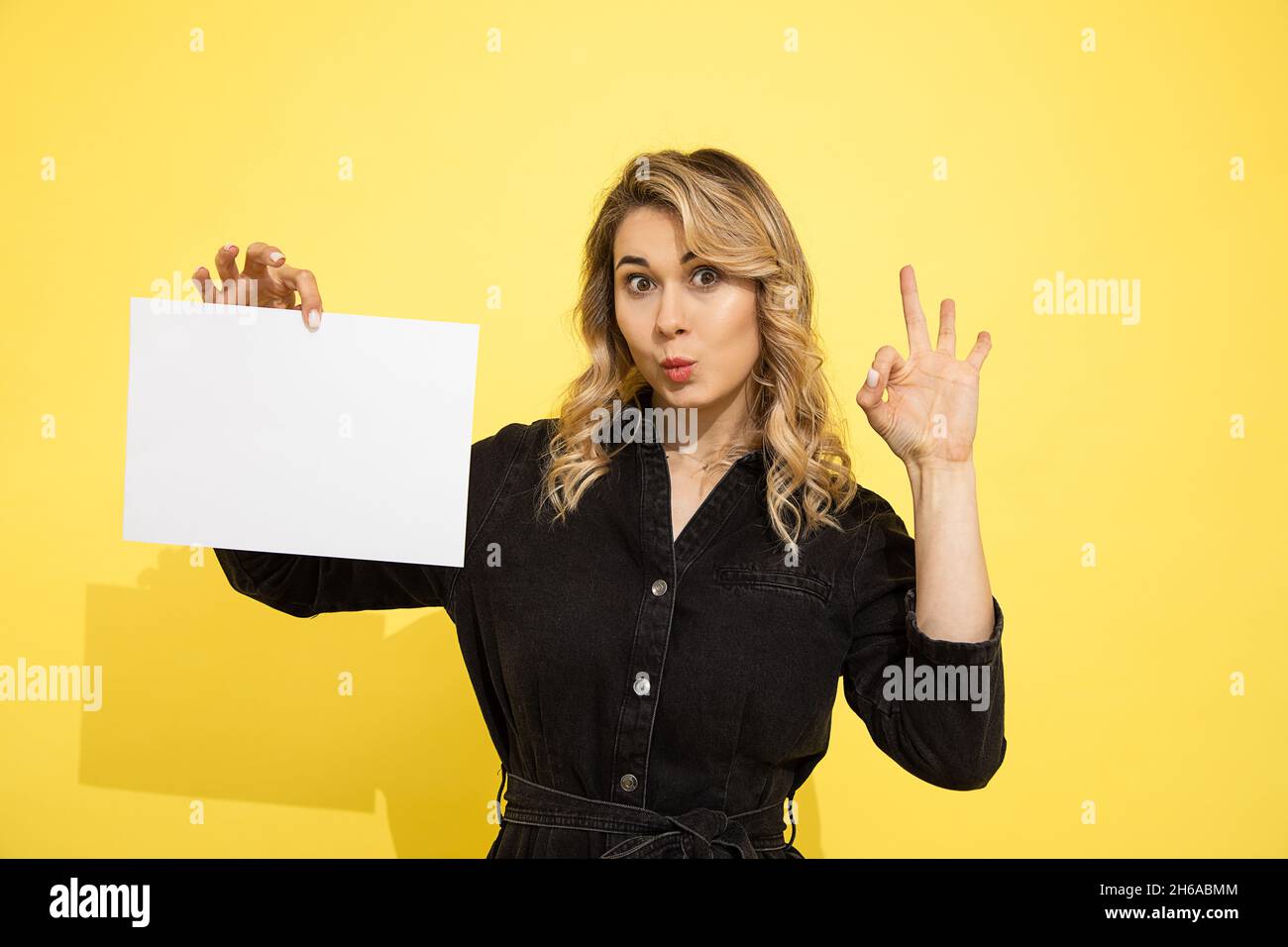 giovane bella donna bionda in abito nero contiene foglio vuoto per l'iscrizione su sfondo giallo in mani puntate con le dita segno ok, sorride e. Foto Stock