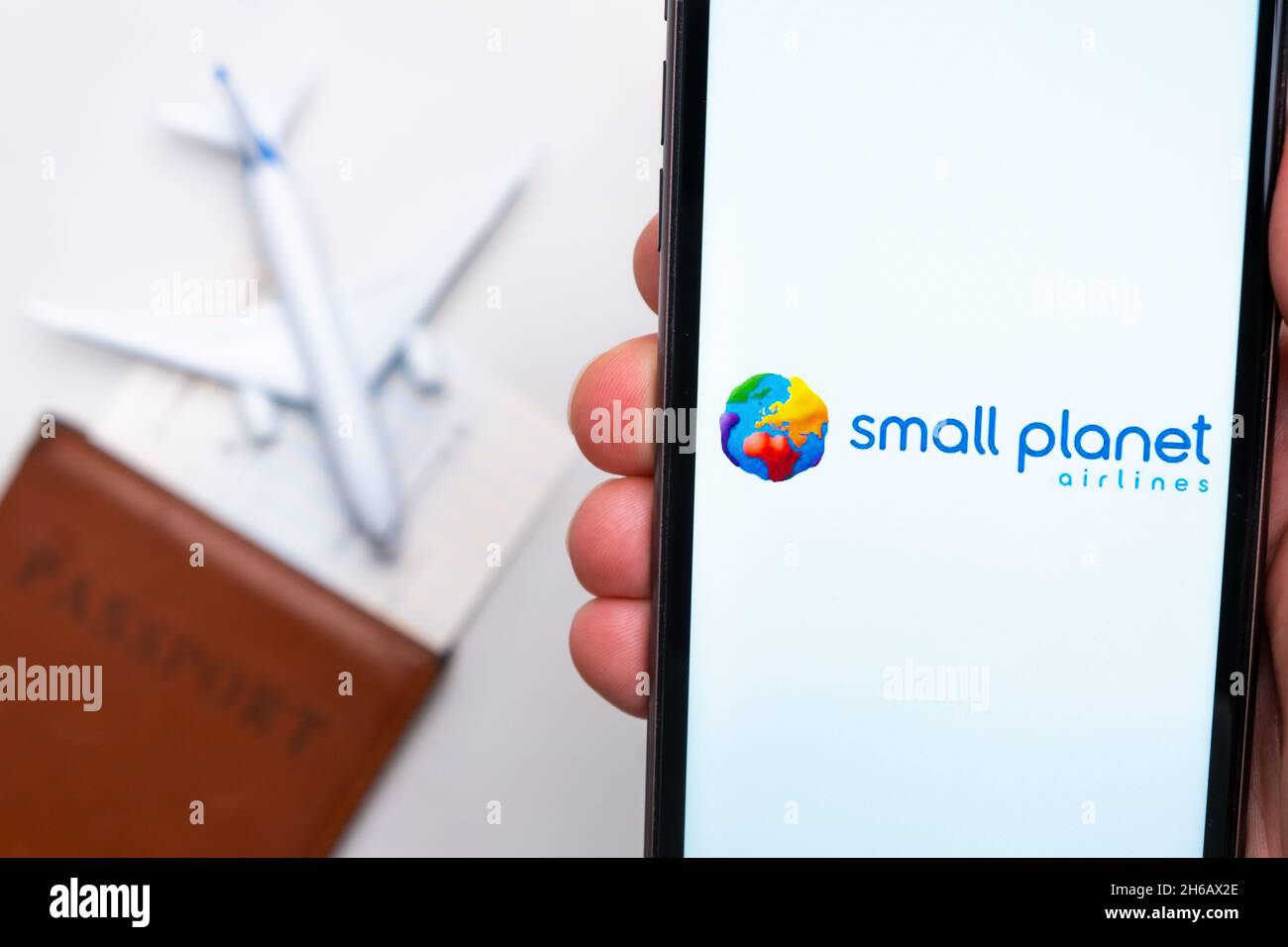 Applicazione Small Planet Airline sullo schermo dello smartphone mans Hand. Un aeroplano giocattolo bianco e un passaporto si trovano su un tavolo con una superficie leggera.Novembre 2021, San Francisco, USA Foto Stock