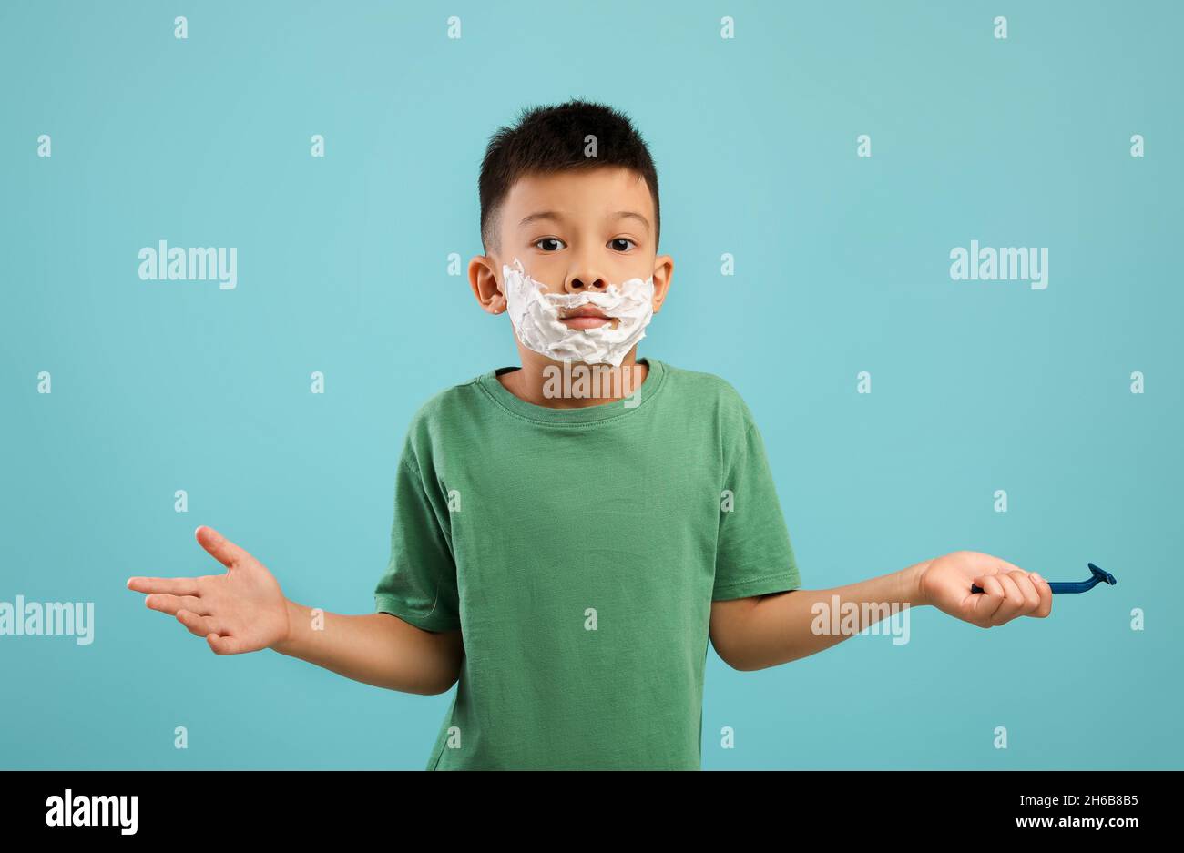 Ritratto di Funny Little Asian Boy con la schiuma da barba sul viso spalle che struggono in confusione, carino coreano Kid Holding Razor e guardando Camer Foto Stock