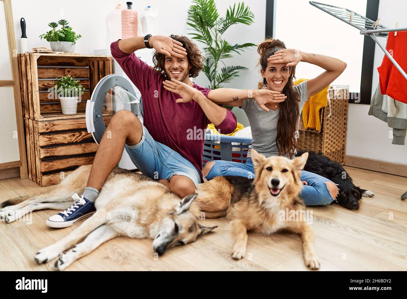 Giovane coppia ispanica che fa lavanderia con cani sorridenti allegri che giocano un boo con le mani che mostrano il volto. Sorpreso ed uscito Foto Stock