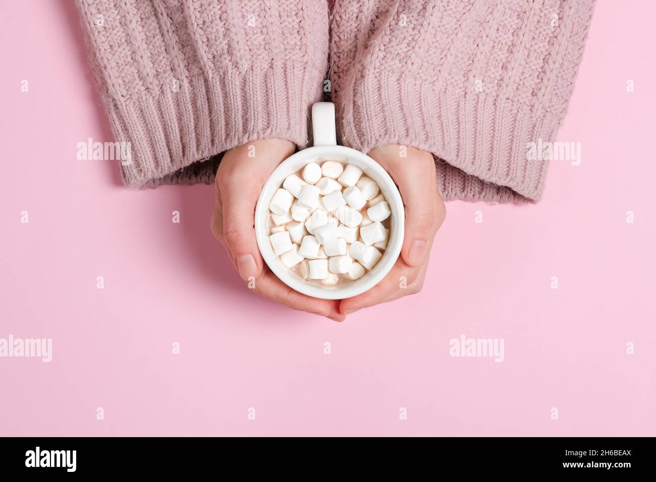 Le ragazze hanno le mani in un pullover color polvere che tiene una grande tazza di cacao o cioccolata calda isolato su sfondo rosa Foto Stock