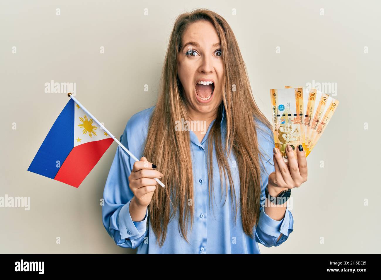 Giovane donna bionda in possesso della bandiera filippina e banconote pesos filippine che celebra pazzo e stupito per il successo con occhi aperti urlando eccitato. Foto Stock