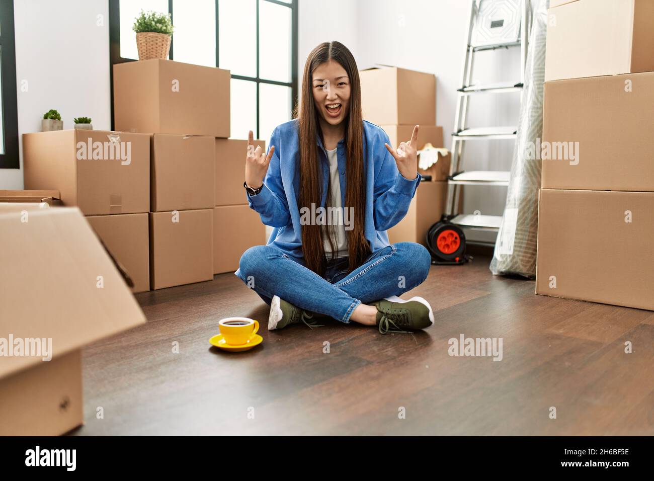 Giovane ragazza cinese seduta sul pavimento a casa nuova gridando con pazza espressione facendo rock simbolo con le mani in su. Star della musica. Concetto pesante. Foto Stock
