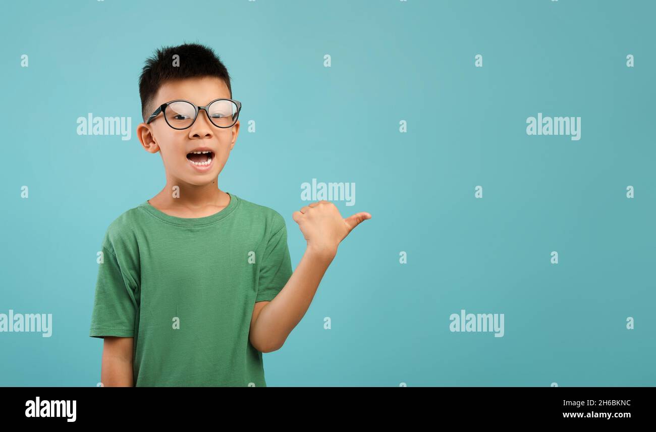 Controllare. Divertente ragazzo asiatico poco in Eyeglasses che punta da parte con Thumb Up, eccitato ragazzo maschio coreano preteen mostrando spazio libero di copia per Advertiseme Foto Stock